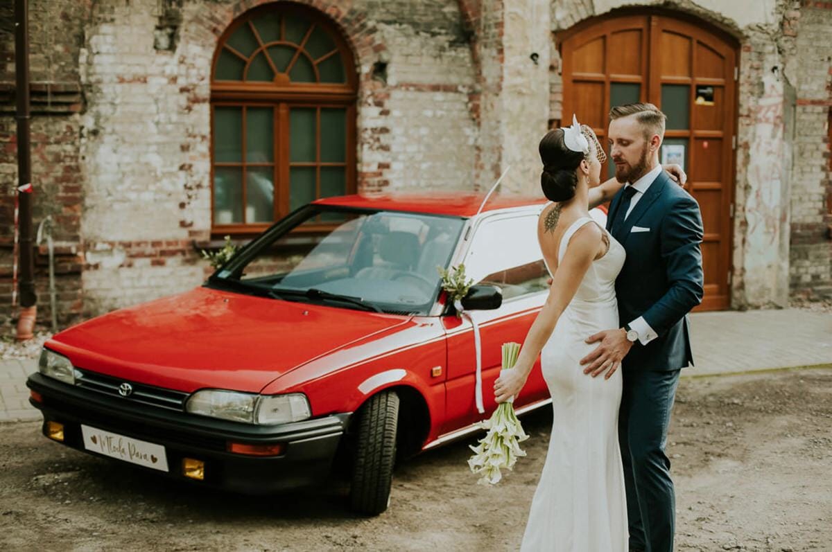 Para młoda przed czerwonym samochodem na tle budynku.