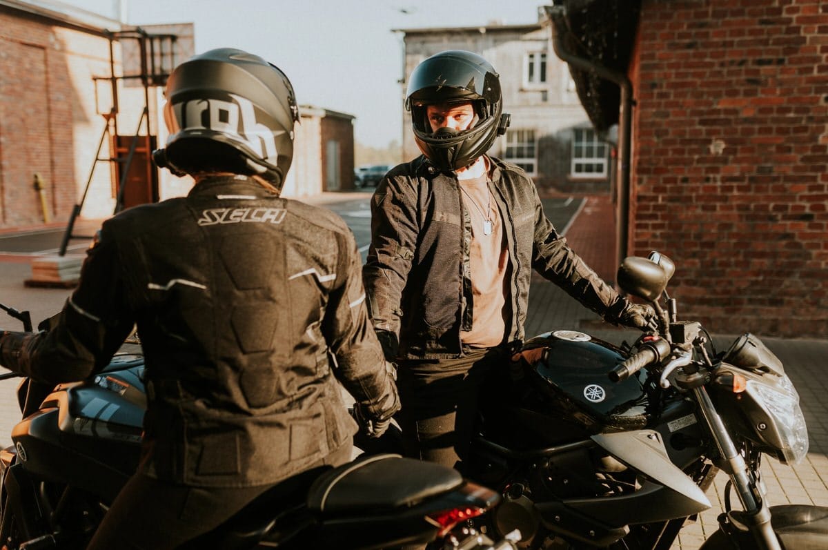 Dwóch motocyklistów w kaskach na parkingu z ceglaną ścianą
