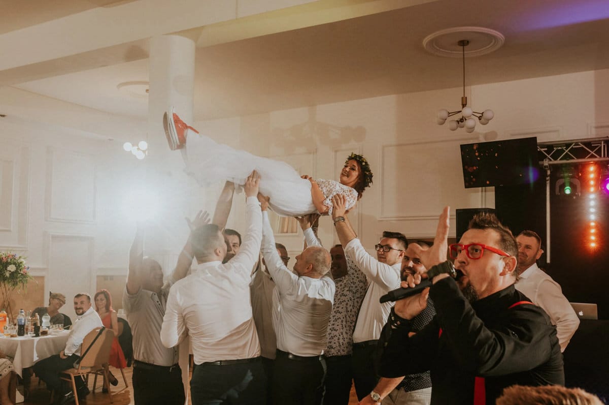 Goście weselni unoszą pannę młodą w białej sukni.